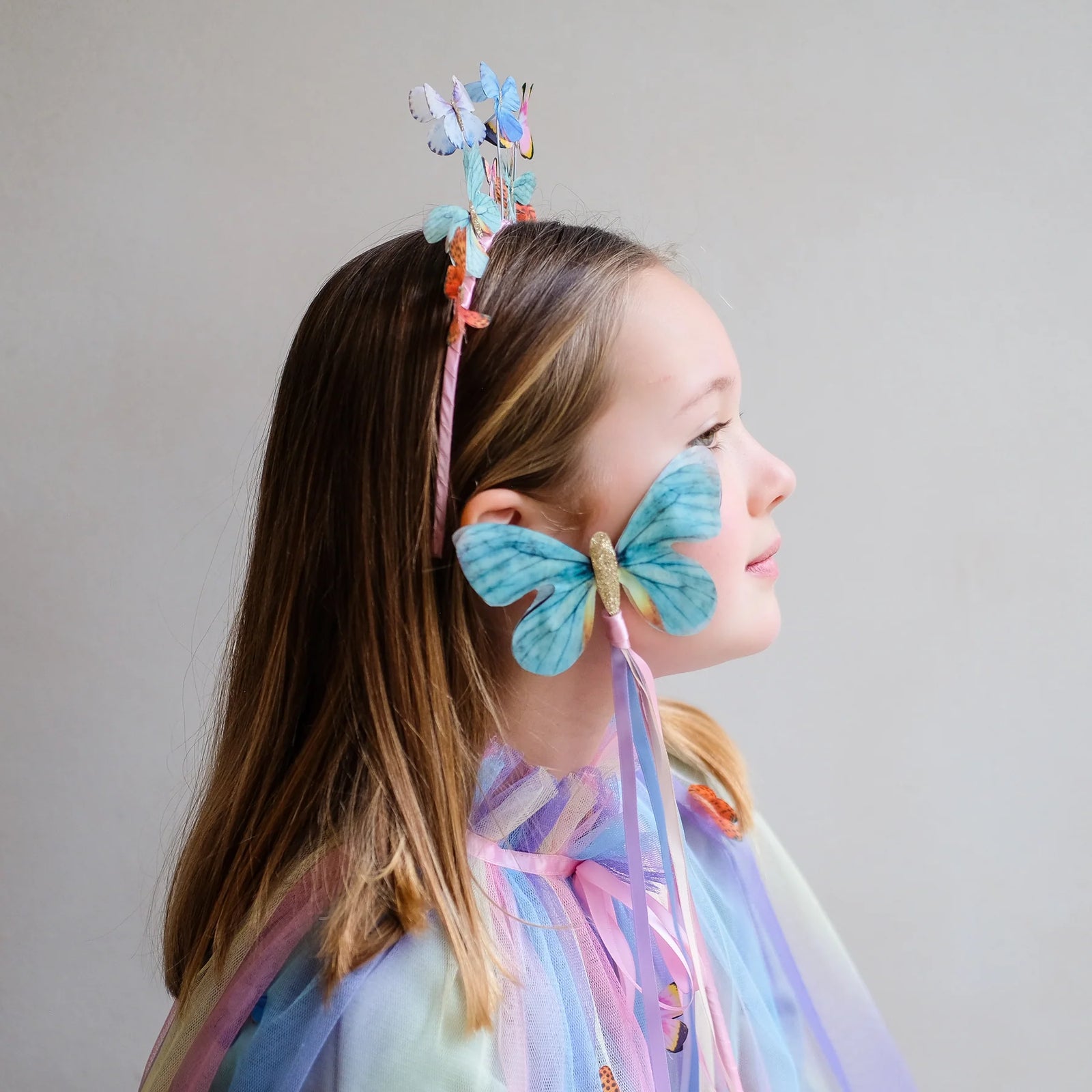 Child in a butterfly costume with a colorful headpiece and wand against a plain background
