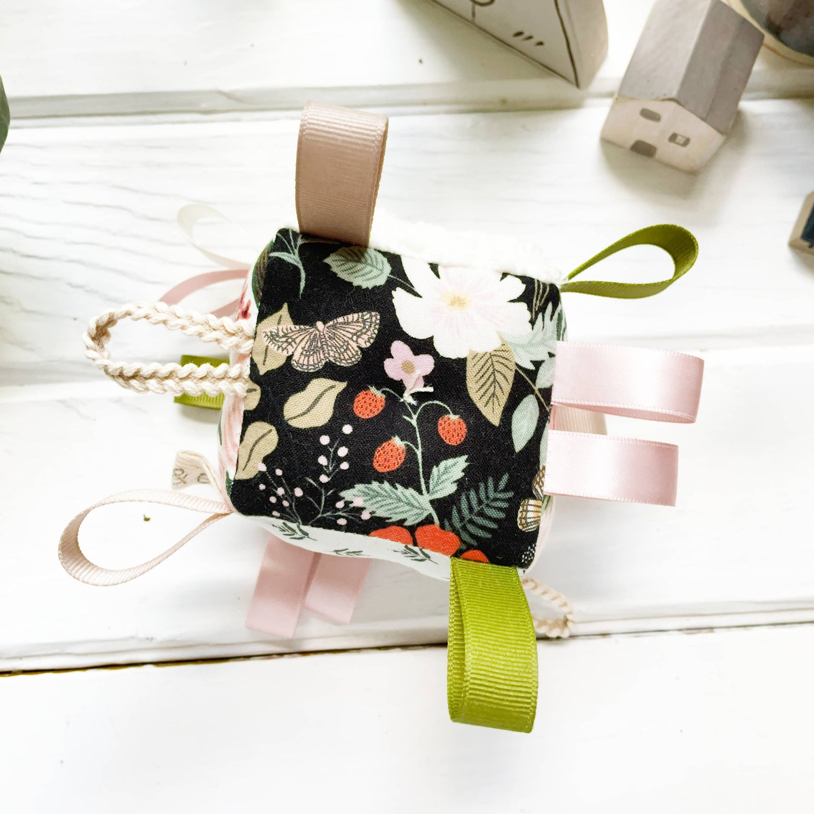 A floral baby rattle block with multiple colors including rose, gold, blush, pale mint, blue, and black, featuring braided loops, ribbons, and a rattle attached to a green ribbon.